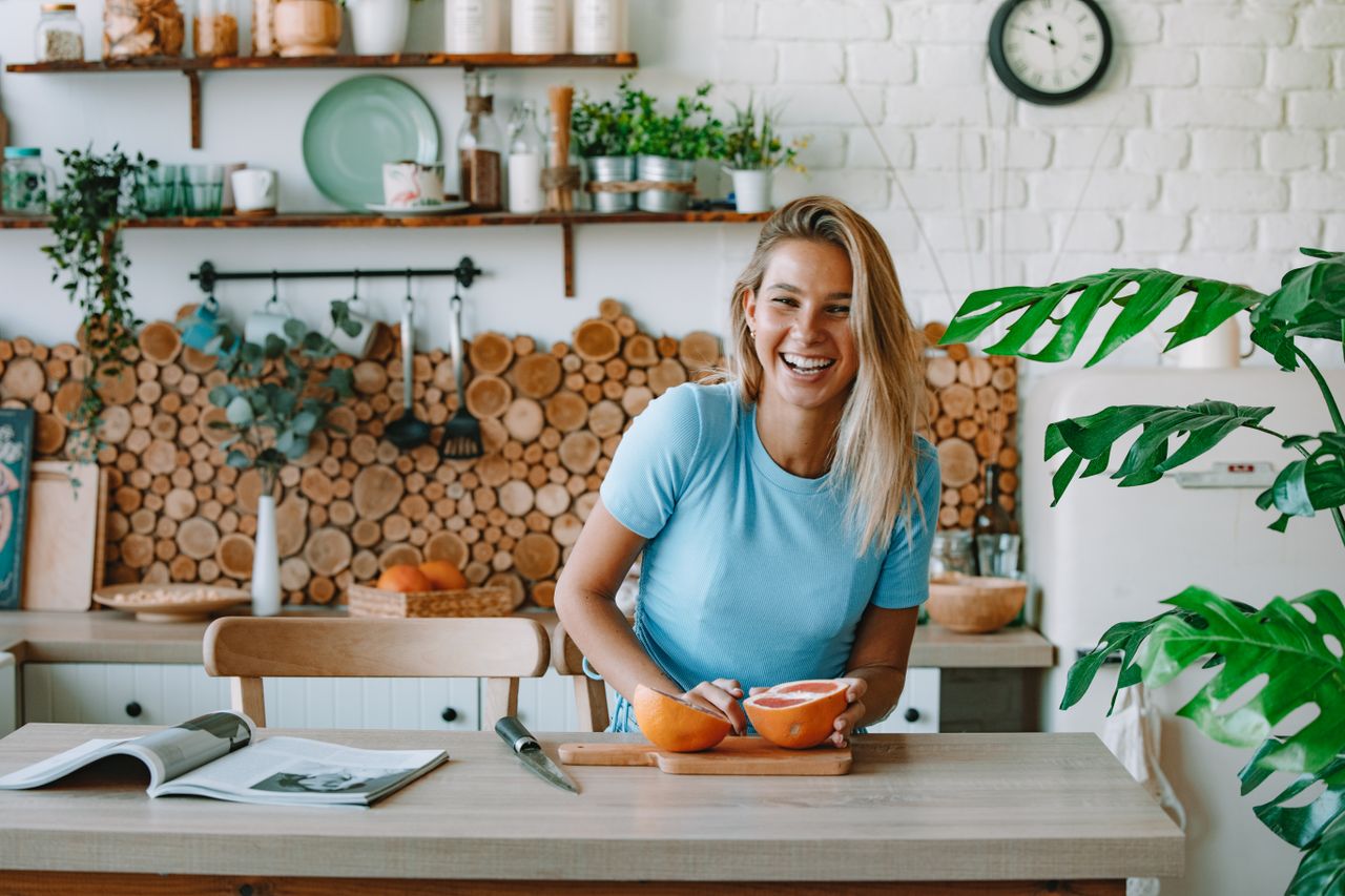 services-03 A joyful woman with blond hair slicing grapefruit in a bright, plant-filled kitchen.
