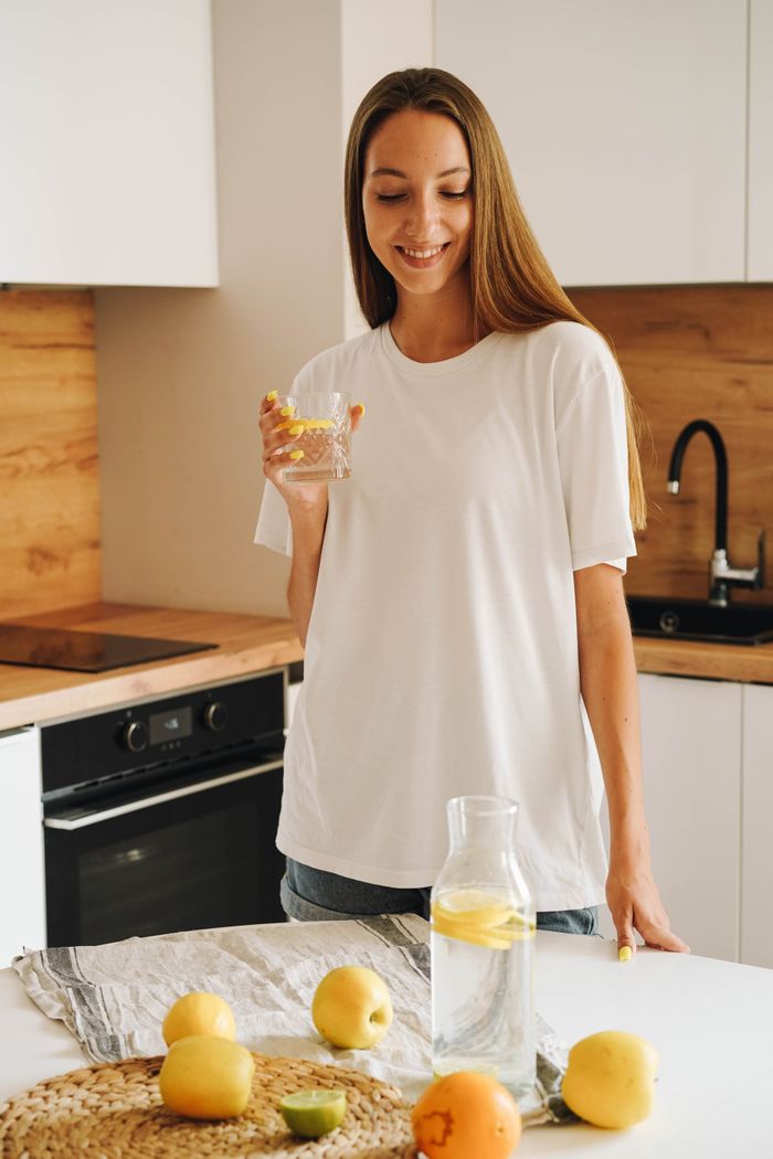 services-04 Smiling woman holding glass of lemon water in a stylish kitchen, promoting a healthy lifestyle.